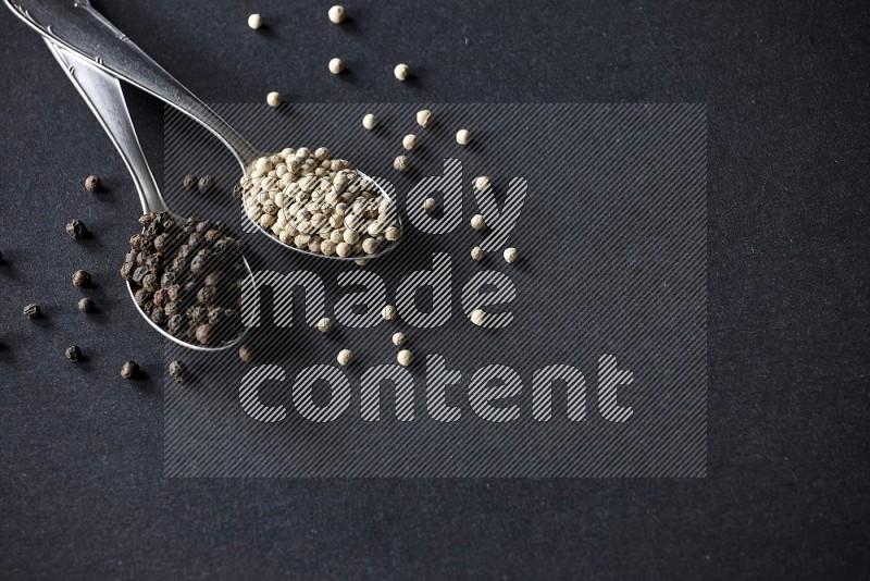 2 metal spoons full of black and white pepper beads with spreaded beads on black flooring