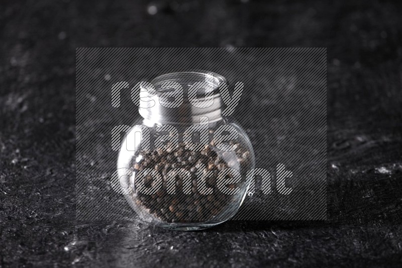 A glass spice jar full of black pepper beads on textured black flooring