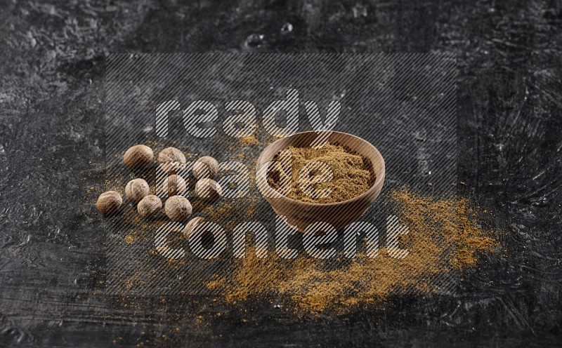 A wooden bowl full of nutmeg powder with whole seeds and sprinkled powder beside it on a textured black flooring