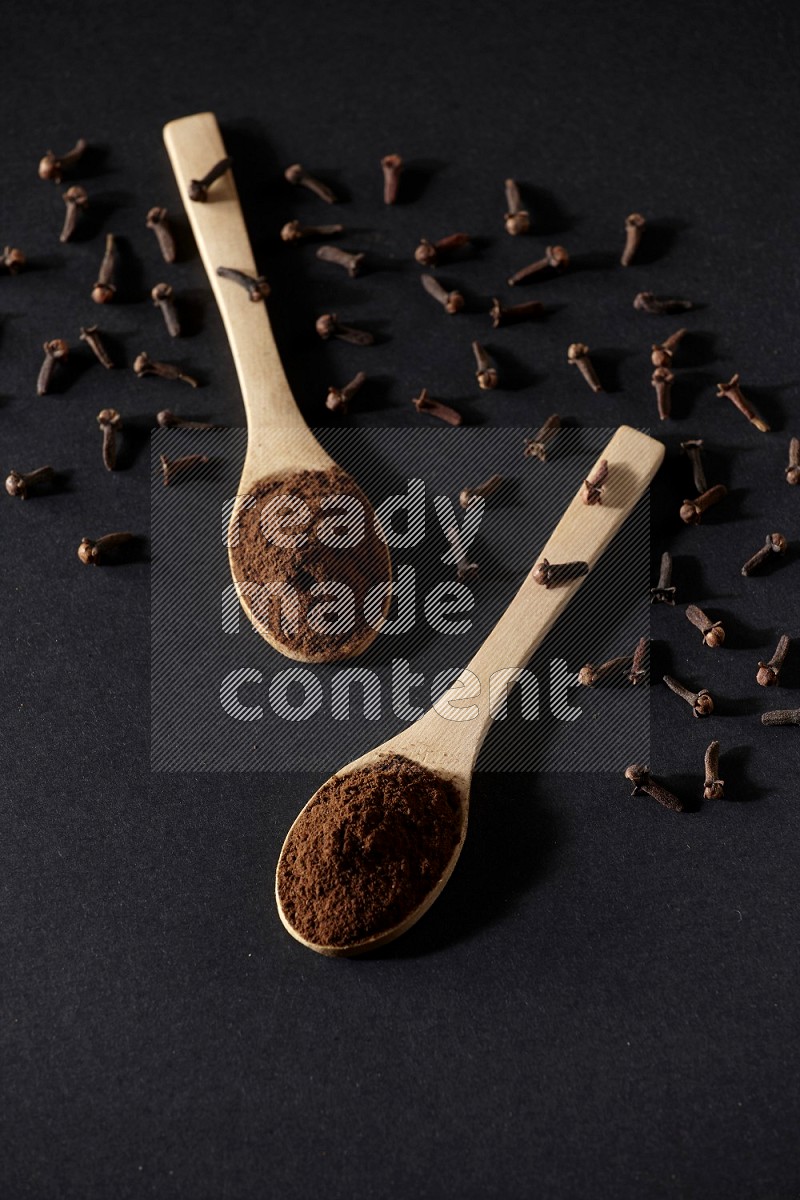 2 wooden spoons full of cloves powder with spreaded whole cloves on a black flooring