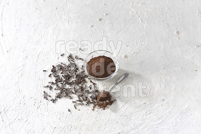 A glass bowl and a metal spoon full of cloves powder with cloves grains spread on a textured white flooring