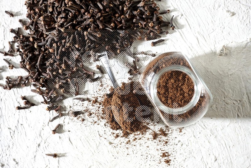 A glass spice jar and a metal spoon full of cloves powder and cloves spread on textured white flooring