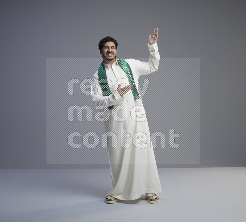 A Saudi man standing wearing thob and Saudi flag scarf on gray background