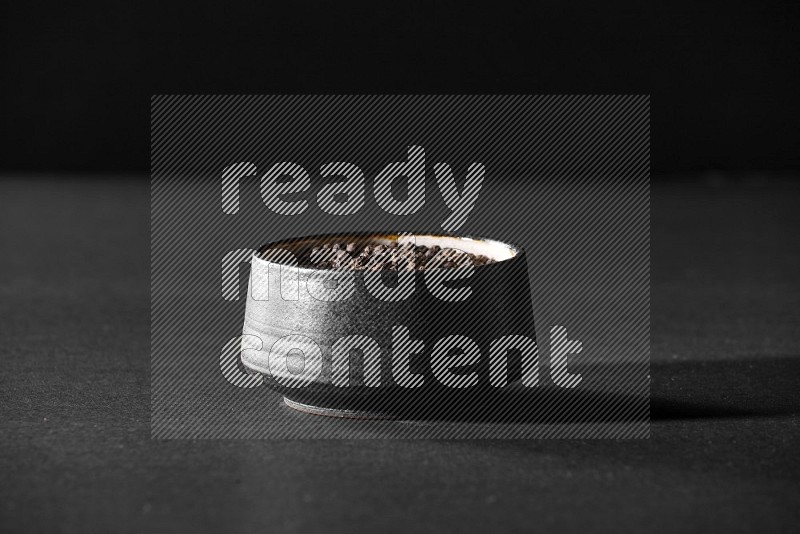 A black pottery bowl full of black pepper beads on black flooring