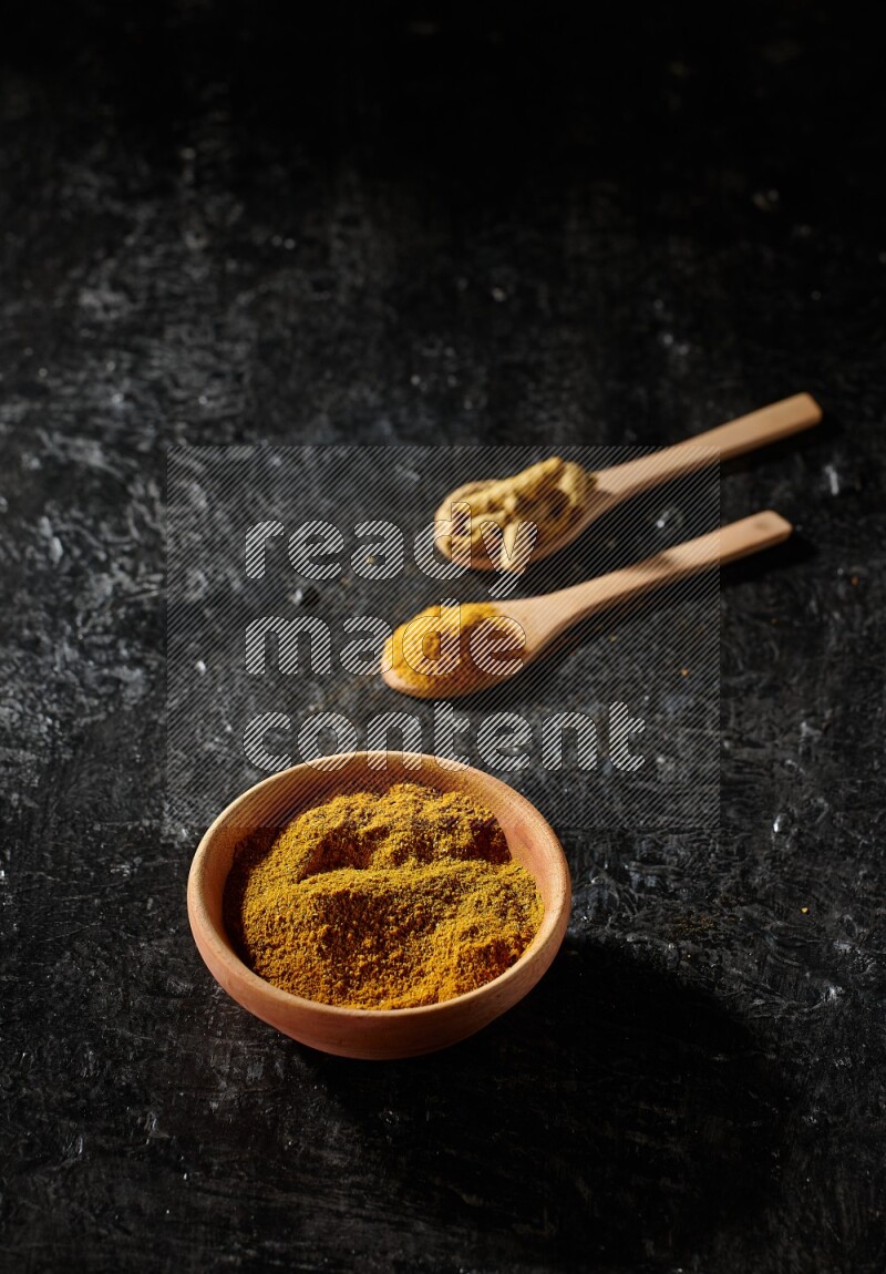 A wooden bowl full of turmeric powder and 2 wooden spoons full of dried turmeric finger and turmeric powder on textured black flooring