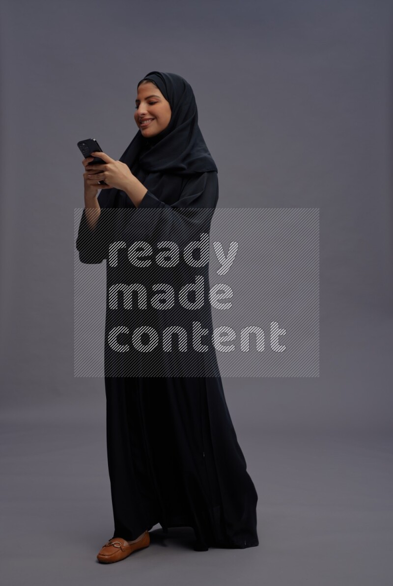 Saudi woman wearing Abaya standing texting on phone on gray background