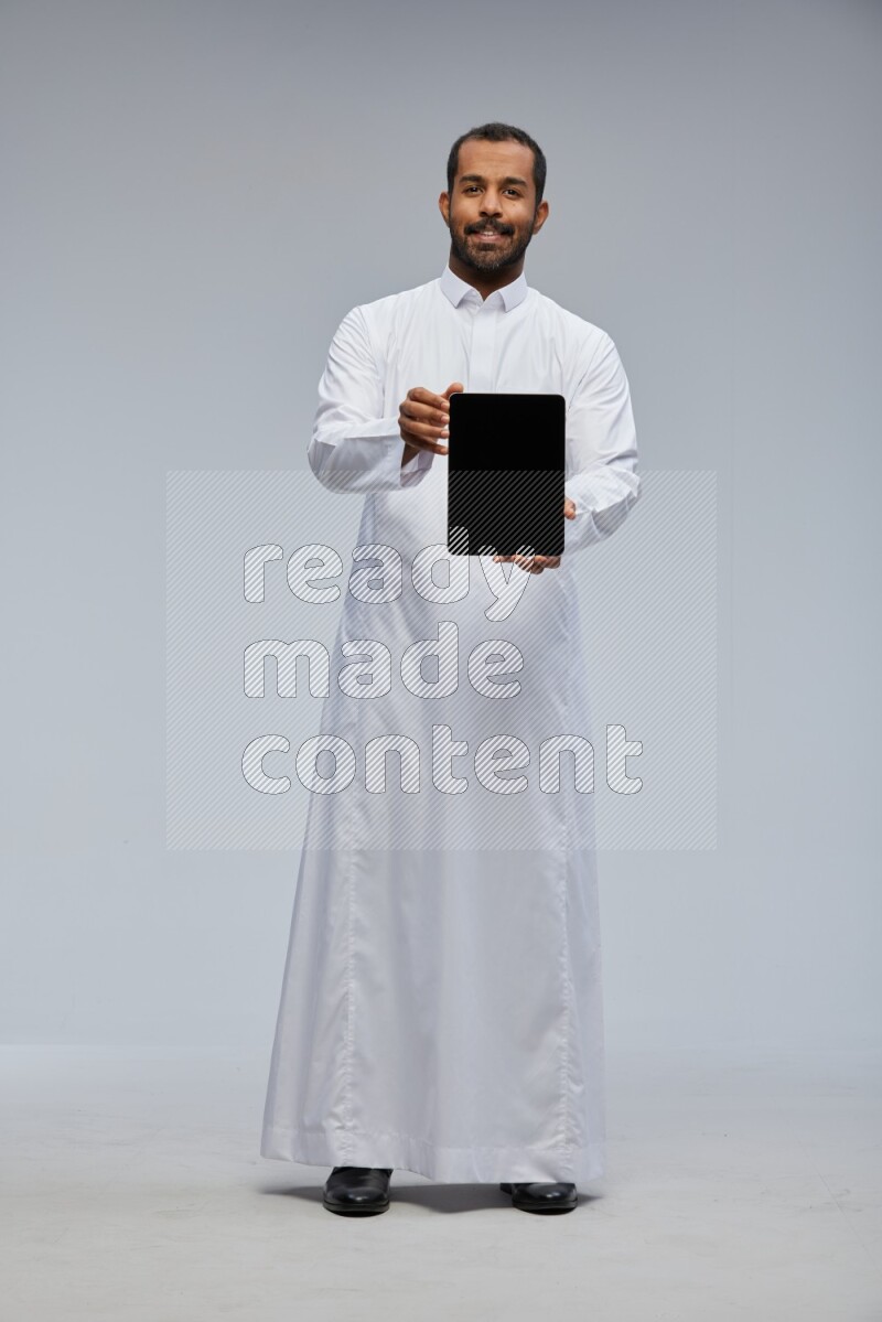 Saudi man wearing Thob standing showing tablet to camera on Gray background