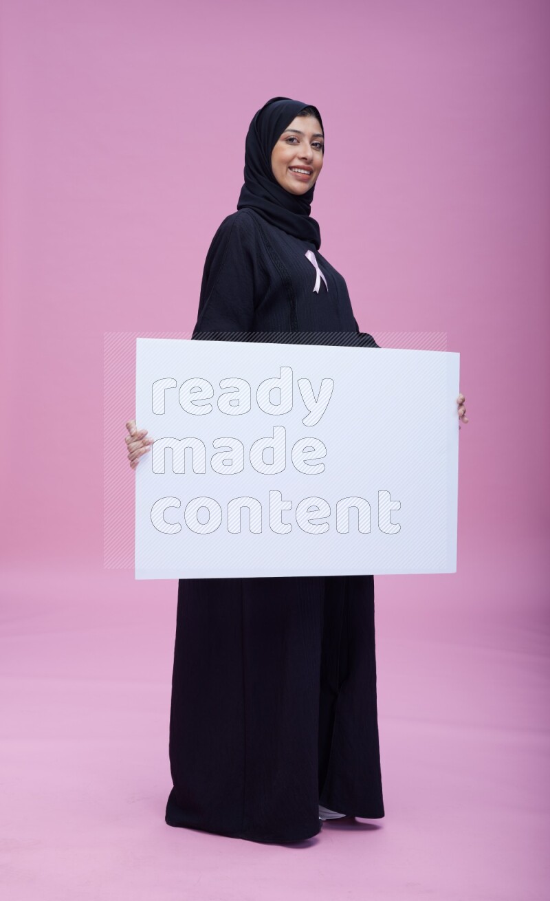 Saudi woman wearing pink ribbon on Abaya standing holding board on pink background