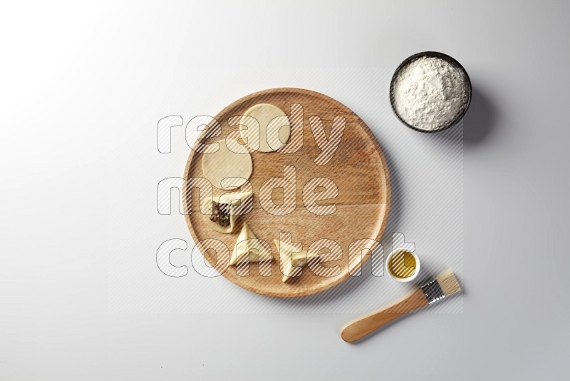 two closed sambosas and one open sambosa filled with meat while flour, and oil with oil brush aside in a wooden dish on a white background