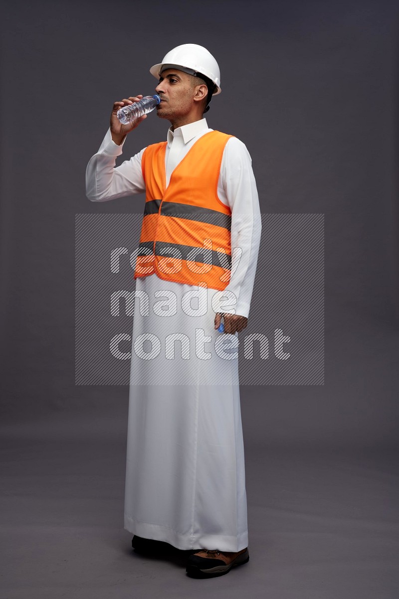 Saudi man wearing thob with engineer vest standing drinking water on gray background
