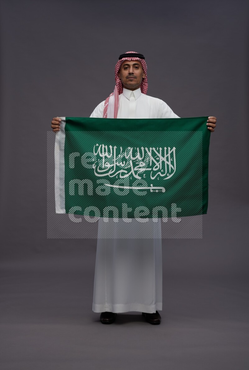 Saudi man wearing thob and shomag standing holding Saudi flag on gray background
