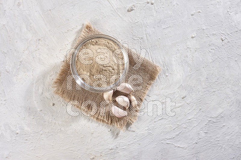 A glass bowl full of garlic powder on burlap fabric with garlic cloves on a textured white flooring in different angles