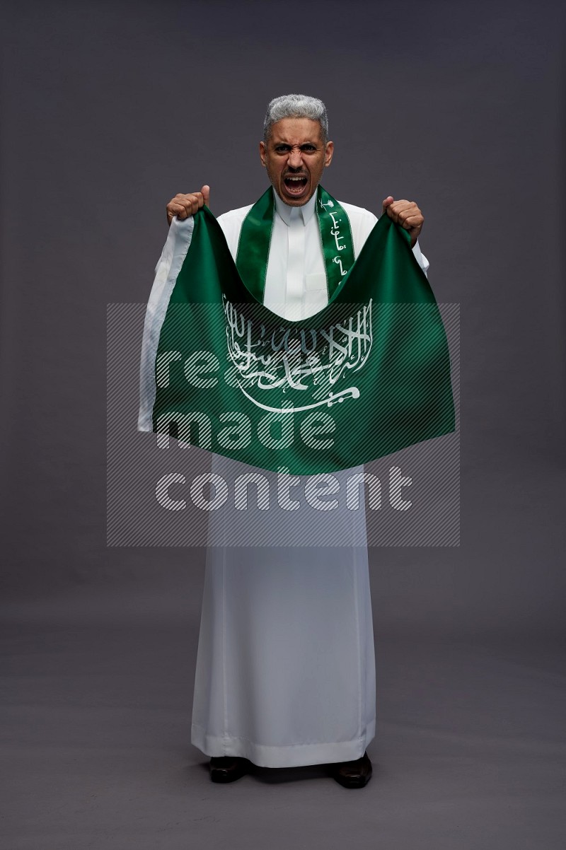 Saudi man wearing thob standing holding Saudi flag on gray background