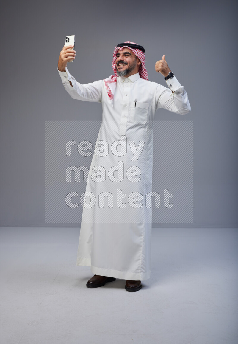 Saudi man Wearing Thob and red Shomag standing taking selfie on Gray background