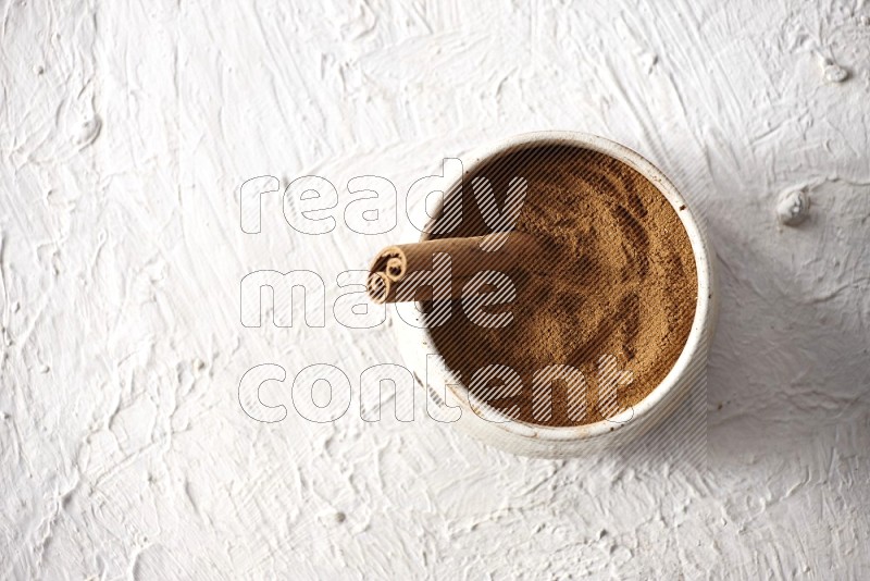 Ceramic beige bowl full of cinnamon powder with a cinnamon stick on a textured white background