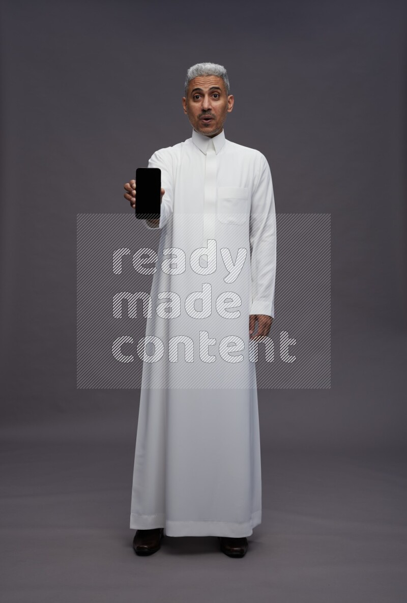 Saudi man wearing thob standing showing phone to camera on gray background