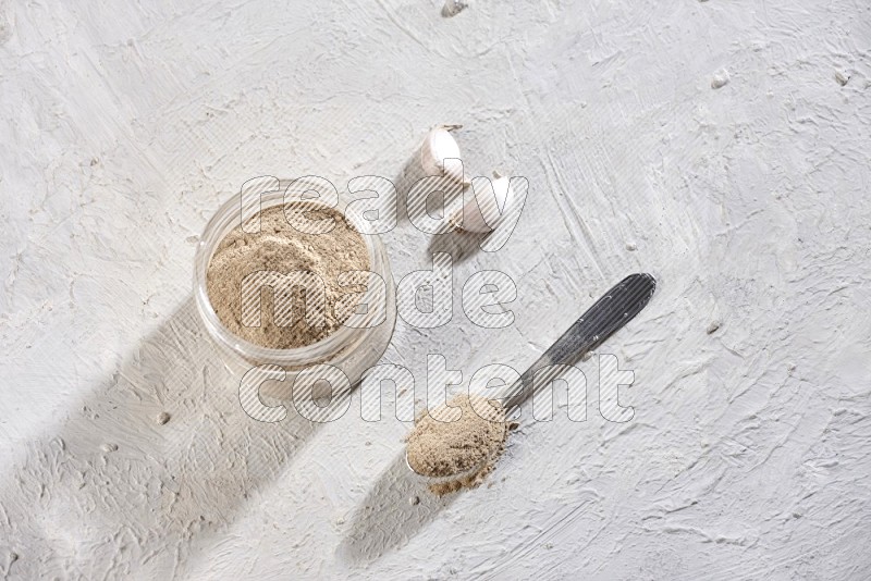 A glass jar full of garlic powder with a metal spoon full of the powder on a textured white flooring