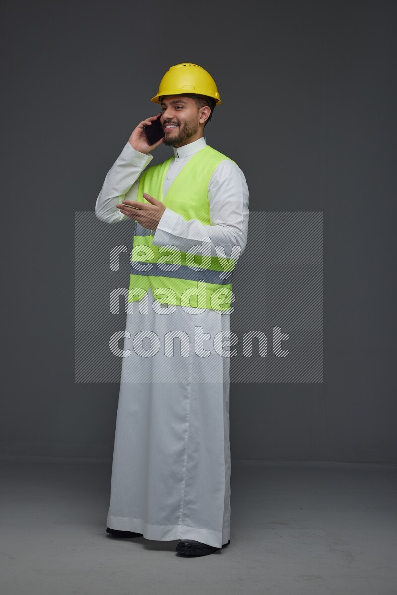 A Saudi man wearing Thobe with a yellow safety vest and white helmet standing and talking in the phone eye level on a gray background