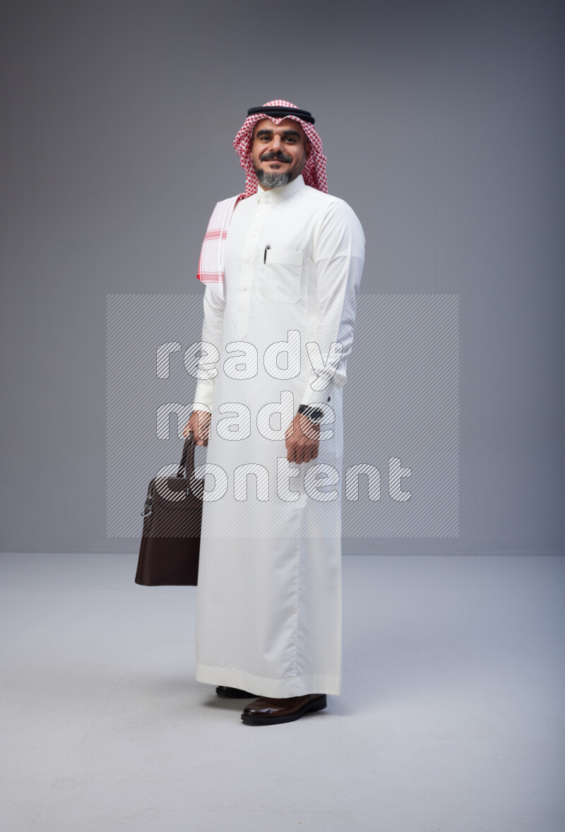 Saudi man Wearing Thob and red Shomag standing holding bag on Gray background