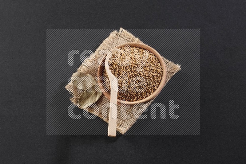 A wooden bowl and spoon full of mustard seeds on a piece of burlap on a black flooring