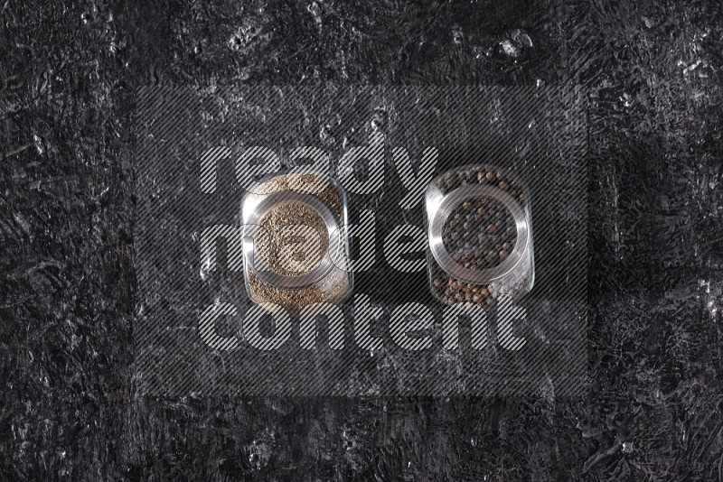 2 glass spice jars full of black pepper powder and black pepper beads on textured black flooring