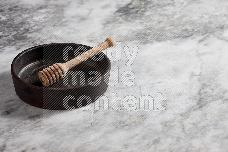Black Pottery Oven Plate with wooden honey handle in it, on grey marble flooring, 45 degree angle