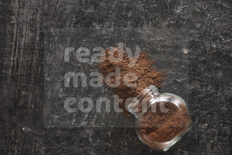 A flipped glass spice jar full of cloves powder and powder came out of it on textured black flooring