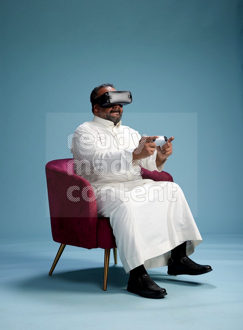 Saudi Man without shimag sitting on chair  gaming with VR and joystick on blue background