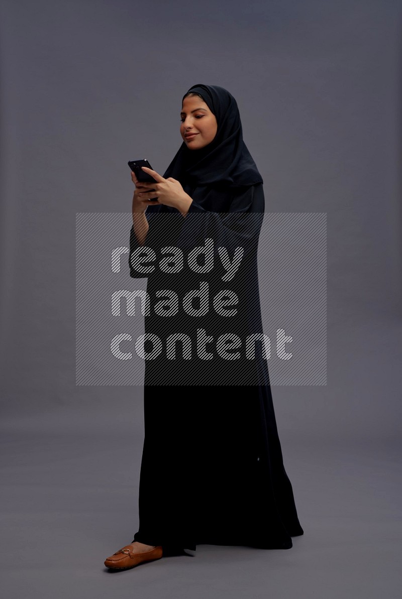 Saudi woman wearing Abaya standing texting on phone on gray background