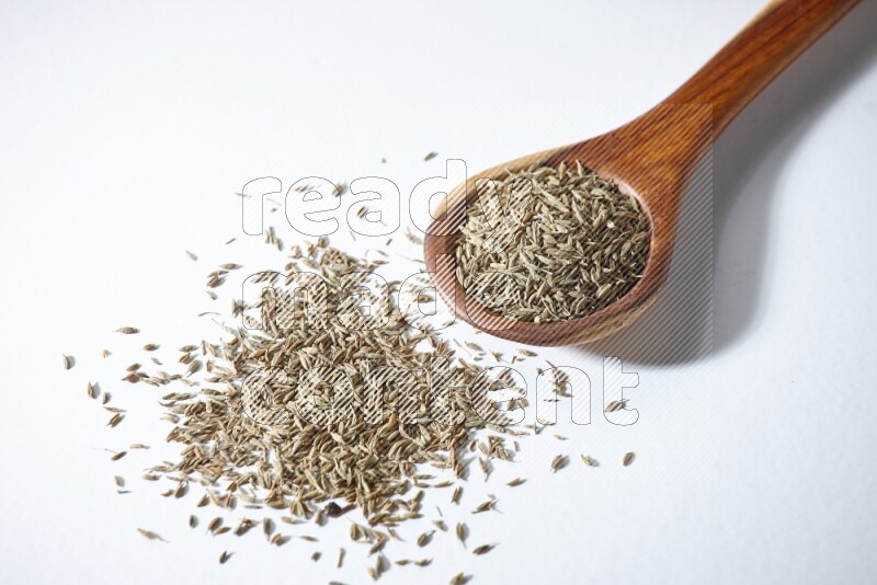 A wooden ladle full of cumin seeds on a white flooring