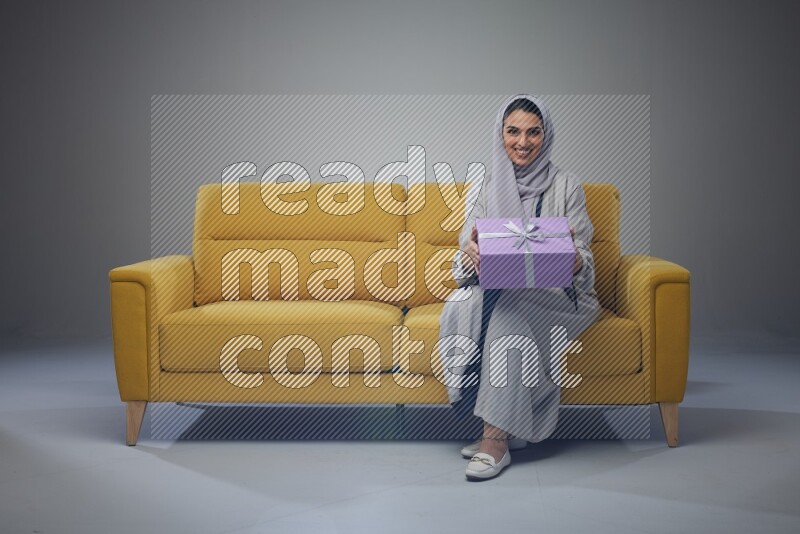 A Saudi woman wearing a light gray Abaya and head scarf sitting on a yellow sofa holding a gift box beside multi color gift boxes eye level on a grey background