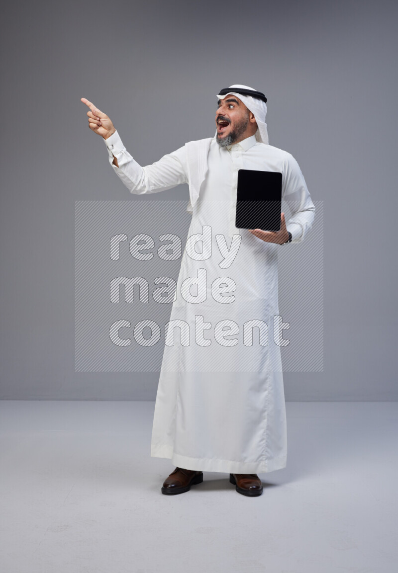 Saudi man Wearing Thob and white Shomag standing showing tablet to camera on Gray background