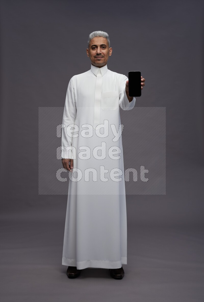 Saudi man wearing thob standing showing phone to camera on gray background