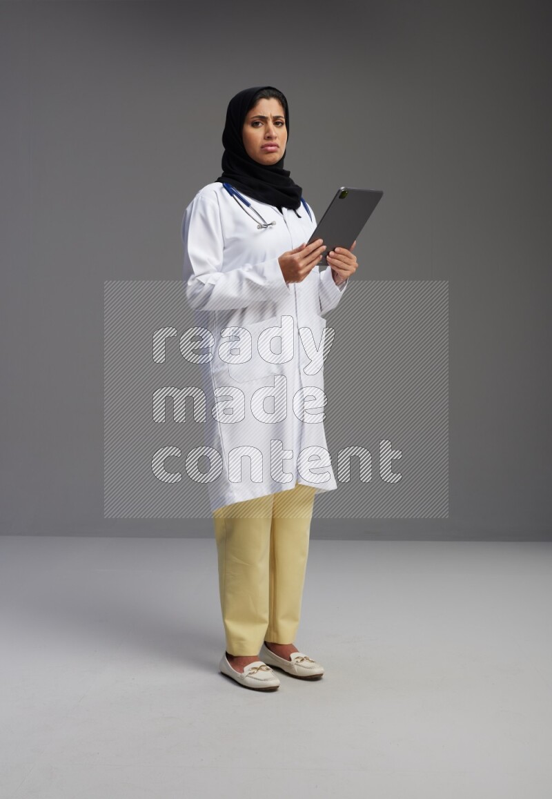 Saudi woman wearing lab coat with stethoscope standing working on tablet on Gray background
