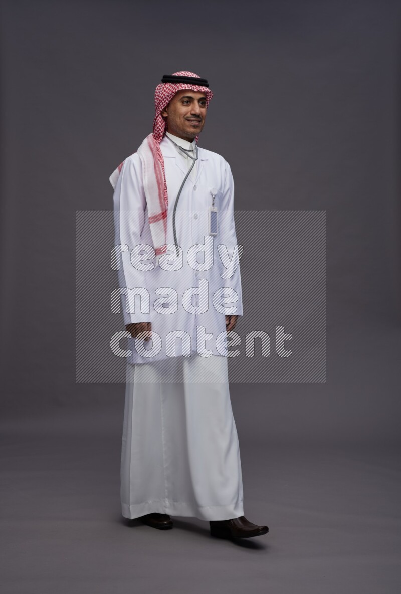 Saudi man wearing thob with lab coat and shomag with pocket employee badge with stethoscope standing interacting with the camera on gray background