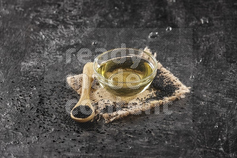 A glass bowl full of black seeds oil and wooden spoon full of black seeds with seeds spreaded on burlap fabric on a textured black flooring