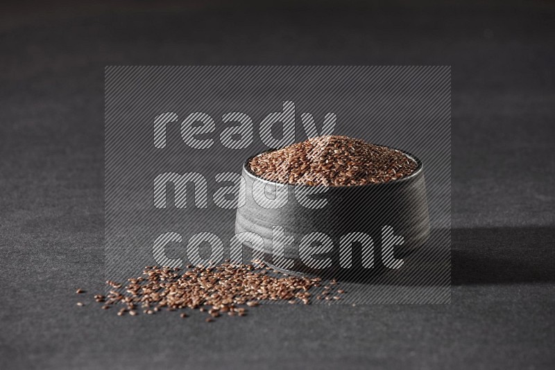 A black pottery bowl full of flax surrounded by the seeds on a black flooring in different angles
