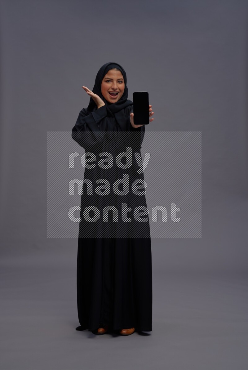 Saudi woman wearing Abaya standing showing phone to camera on gray background