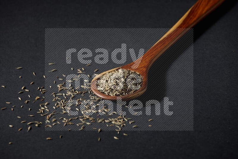 A wooden ladle full of cumin seeds on black flooring