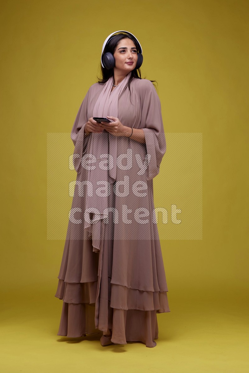 A Saudi woman Listing to Music on a Yellow Background wearing Brown Abaya