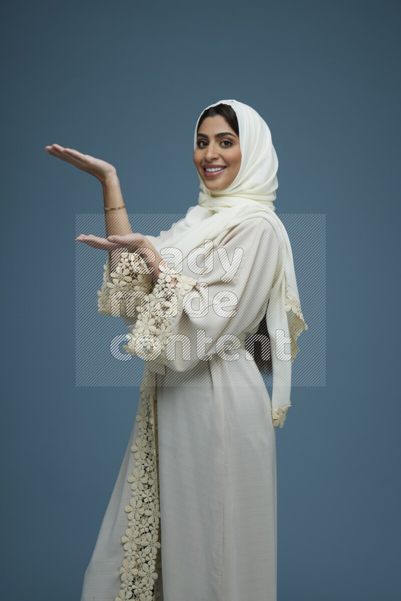 A Saudi woman pointing in a blue background wearing an off-white Abaya Hijab