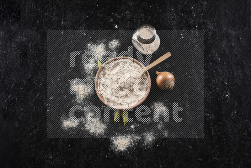 A wooden bowl full of onion powder with a glass jar beside it and fresh onion on black background