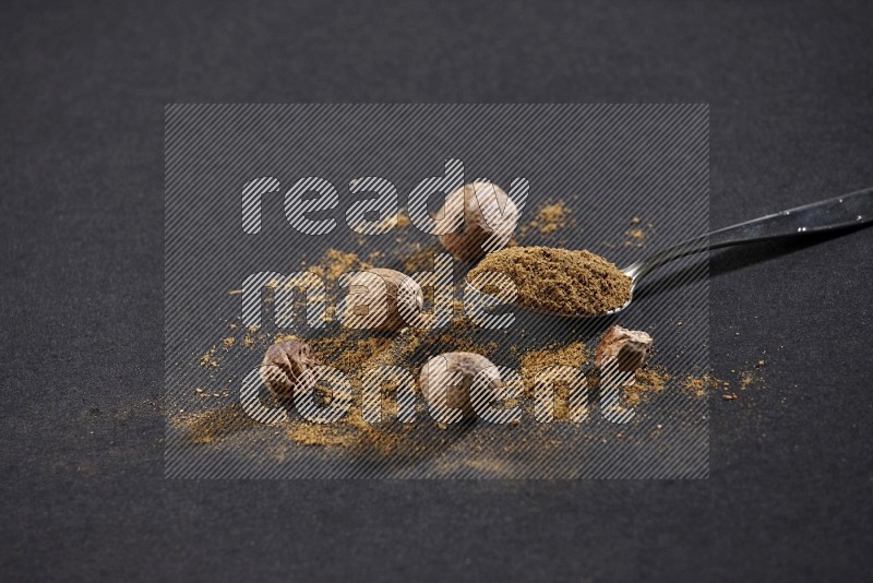 A metal spoon full of nutmeg powder with nutmeg powder and seeds beside it on a black flooring in different angles