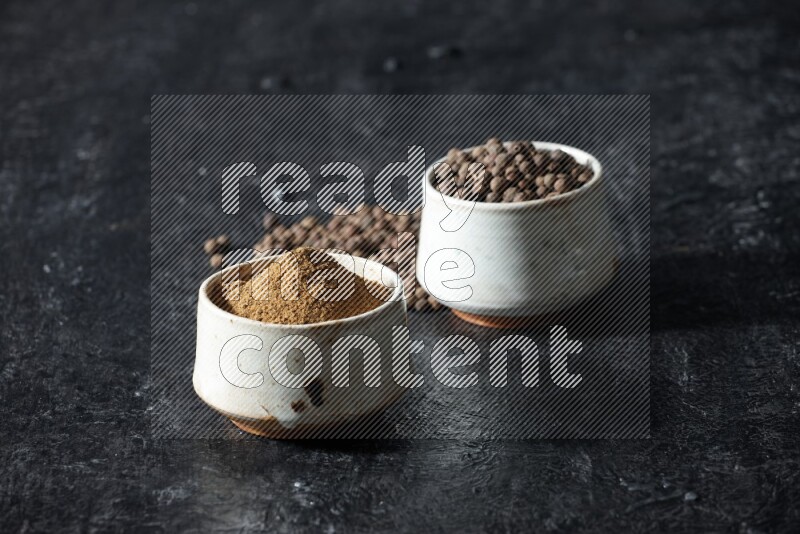 2 beige bowls, one full of allspice powder and the other full of whole balls and both spread on a textured black flooring