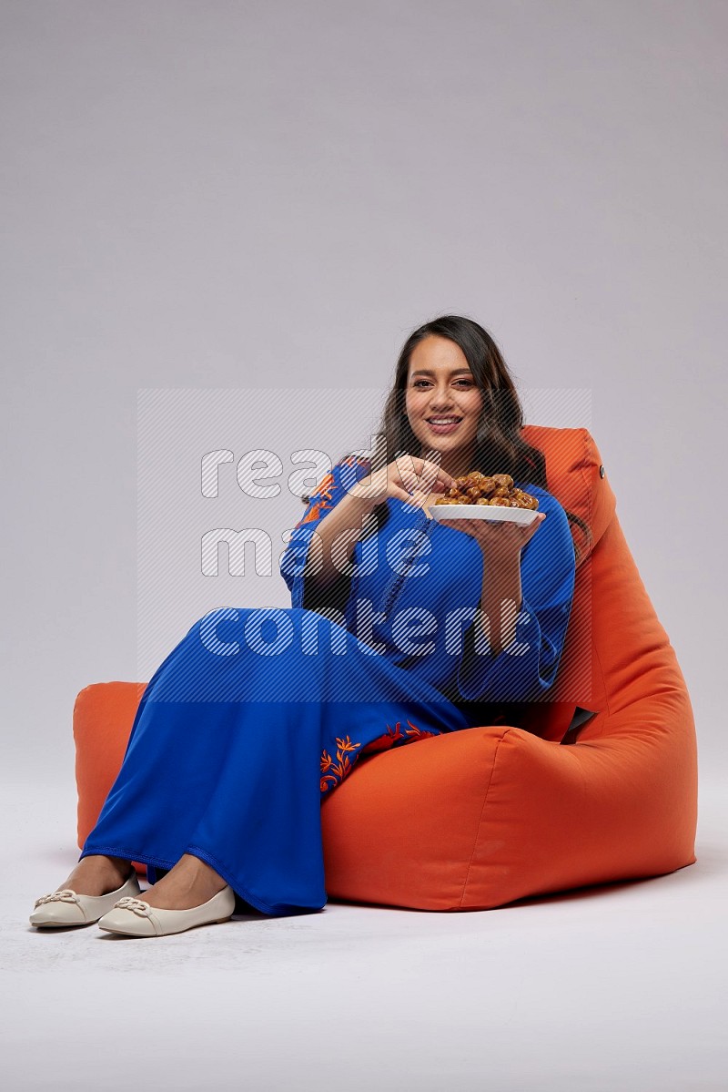 A Woman sitting on an orange beanbag wearing Jalabeya holding a plate of dates