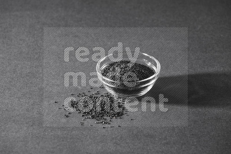 A glass bowl full of black seeds and seeds spreaded beside it on a black flooring