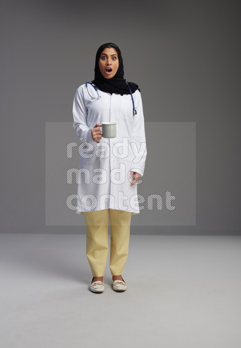 Saudi woman wearing lab coat with stethoscope standing holding mug on Gray background