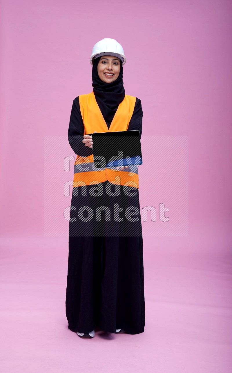 Saudi woman wearing Abaya with engineer vest and helmet standing showing tablet to camera on pink background