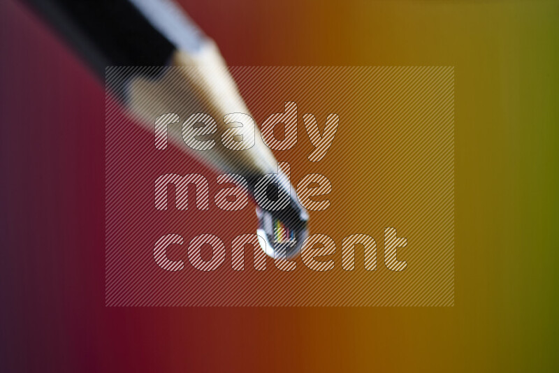 The image captures a close-up of a water droplet balanced on the sharp tip of a pencil, set against a blurred colorful background