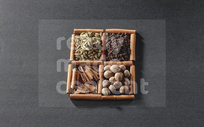 4 مربعات من عيدان القرفة ممتلئة بقطع القرفة وحبوب الهيل والقرنفل وجوزة الطيب على أرضية سوداء